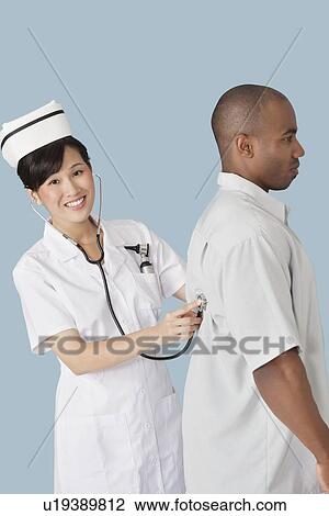 Stock Image - Portrait of a happy female doctor examining male patient's back with stethoscope over light blue background. Fotosearch