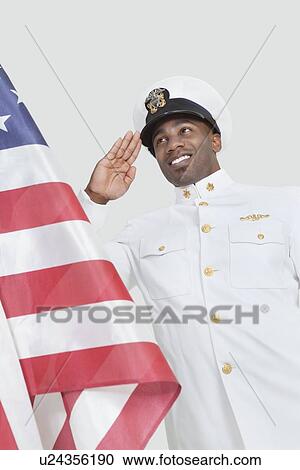 Stock Image - Portrait of a happy young US Navy officer saluting American flag over gray background. Fotosearch