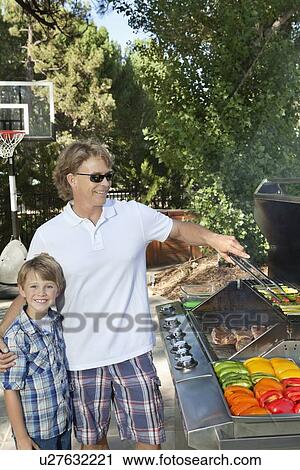 Stock Image - Portrait of a little boy with father barbecuing vegetable on barbecue grill in lawn. Fotosearch