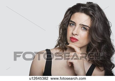 Stock Image - Portrait of a thoughtful young woman with hand on chin over gray background. Fotosearch
