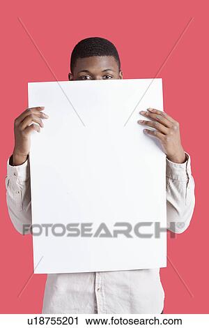 Stock Image - Portrait of a young man holding blank cardboard in front of face over pink background. Fotosearch