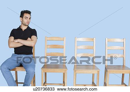 Portrait of a young man sitting beside empty wooden chairs over blue background View Large Photo Image Stock Image - Portrait of a young man sitting beside empty wooden chairs over blue background. Fotosearch