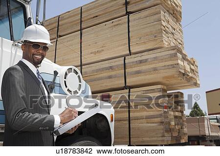 Stock Image - Portrait of African American male contractor writing notes while standing by logging truck. Fotosearch