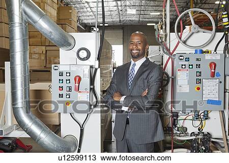 Stock Image - Portrait of African American businessman standing arms crossed with machinery in background. Fotosearch