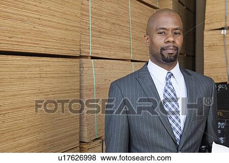 Stock Photo - Portrait of an African American businessman standing in front of stacked wooden planks. Fotosearch