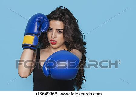 Stock Photo - Portrait of an aggressive young woman wearing boxing gloves over blue background. Fotosearch