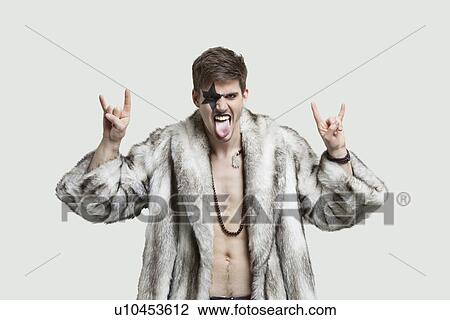 Stock Image - Portrait of an angry young man making rebellious gestures and sticking out tongue against gray background. Fotosearch
