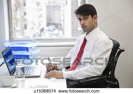 Picture - Portrait of an Indian businessman writing on paper at desk in office. Fotosearch