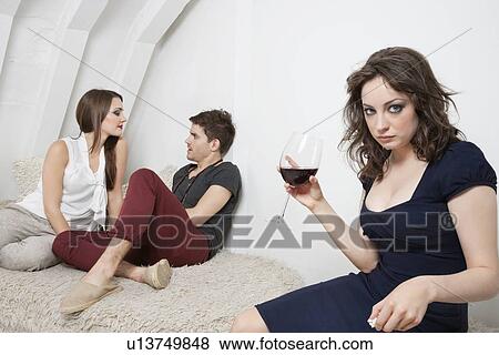 Stock Photo - Portrait of an irritated young woman holding wine glass with couple in the background. Fotosearch