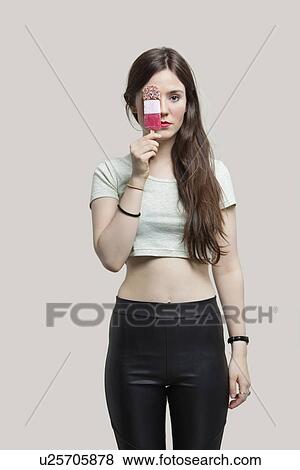 Stock Photo - Portrait of beautiful young woman holding ice cream bar over eye against gray background. Fotosearch