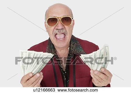 Stock Image - Portrait of excited senior man showing US banknotes with mouth open against gray background. Fotosearch