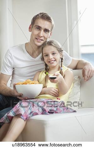 Picture - Portrait of father and daughter watching TV with bowl full of wheel shape snack pellets. Fotosearch