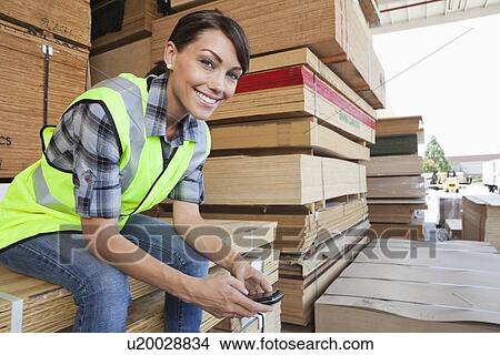 Picture - Portrait of female industrial worker using cell phone while sitting on stack of wooden planks. Fotosearch