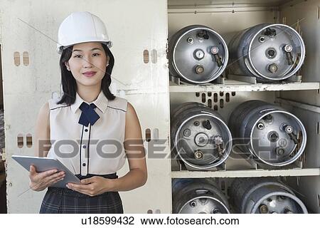 Stock Image - Portrait of female industrial worker holding tablet PC with cylinders on shelf in background. Fotosearch