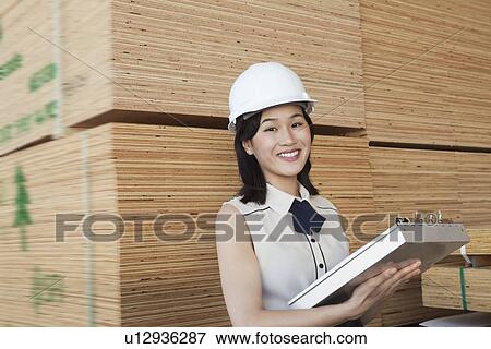 Stock Photo - Portrait of female industrial worker holding clipboard with stacked wooden planks in background. Fotosearch
