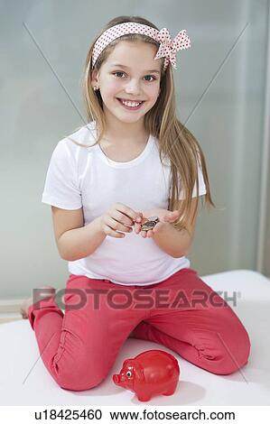 Stock Image - Portrait of girl with piggy bank and coins sitting in bed. Fotosearch
