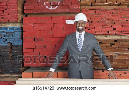 Stock Image - Portrait of happy African American male contractor standing in front of stacked wooden planks. Fotosearch