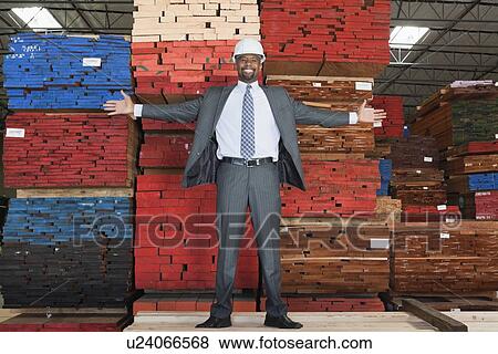 Stock Photo - Portrait of happy African American male engineer standing with arms outstretched in front of stacked wooden planks. Fotosearch