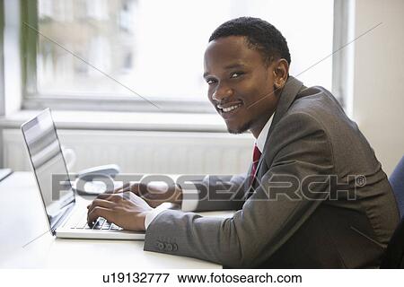 Stock Photo - Portrait of happy African American businessman using laptop at office desk. Fotosearch