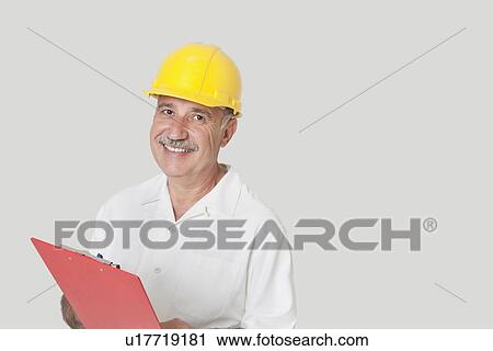 Stock Image - Portrait of happy senior constructor with clipboard over gray background. Fotosearch