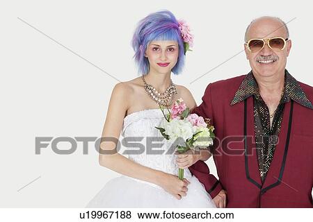 Stock Photo - Portrait of happy senior man standing arm in arm with beautiful daughter in wedding dress against gray background. Fotosearch