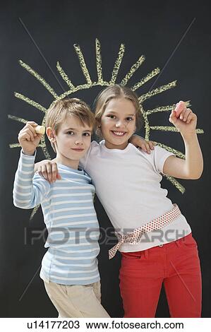 Stock Image - Portrait of happy siblings holding chalks against blackboard. Fotosearch