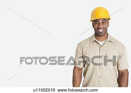 Stock Photo - Portrait of happy young African man wearing yellow hard hat helmet over gray background. Fotosearch