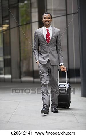 Stock Image - Portrait of happy young businessman walking with wheeled suitcase. Fotosearch