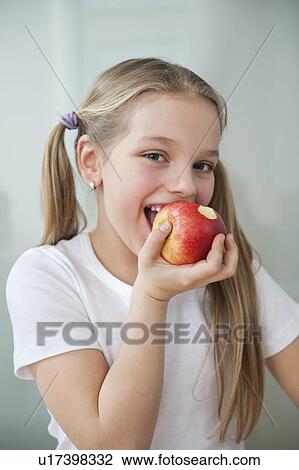 Stock Image - Portrait of happy young girl eating an apple over gray background. Fotosearch