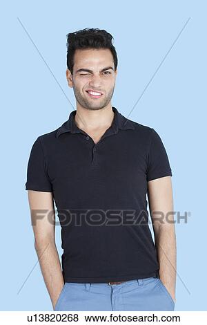 Stock Photo - Portrait of mid adult man winking with hands in pockets over blue background. Fotosearch