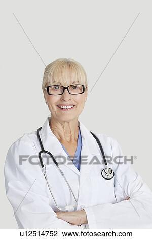 Stock Image - Portrait of senior female doctor with arms crossed over gray background. Fotosearch