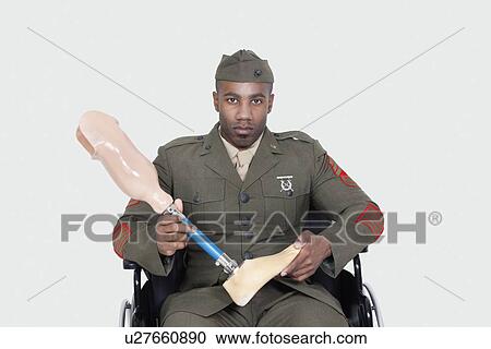 Stock Image - Portrait of US military officer in wheelchair holding prosthesis foot over gray background. Fotosearch