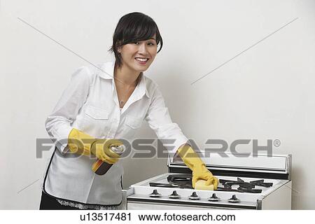 Stock Image - Portrait of young housemaid cleaning stove against gray background. Fotosearch