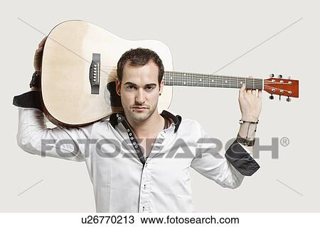 Stock Image - Portrait of young man carrying guitar over his shoulder against gray background. Fotosearch