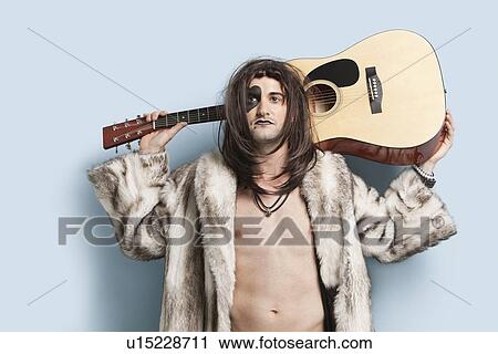 Stock Image - Portrait of young man holding guitar behind back against light blue background. Fotosearch