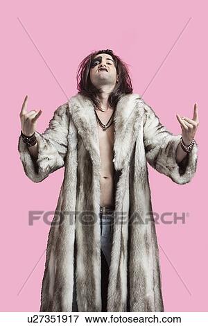 Stock Photo - Portrait of young man in fur coat gesturing rock music sign over pink background. Fotosearch