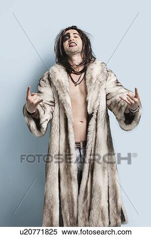 Stock Photography - Portrait of young man in fur coat gesturing rock music sign against light blue background. Fotosearch