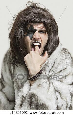 Stock Image - Portrait of young man in fur coat with finger in mouth against gray background. Fotosearch