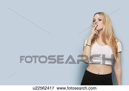 Stock Photo - Portrait of young woman smoking cigarette against light blue background. Fotosearch