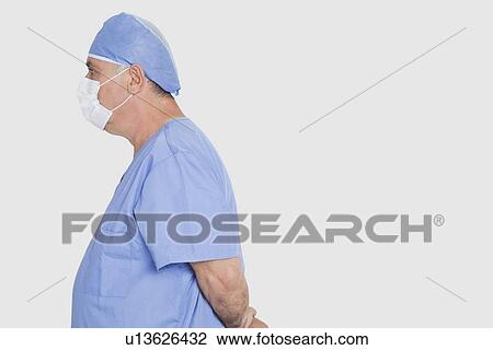 Stock Image - Profile shot of senior male surgeon standing with hands behind back over gray background. Fotosearch