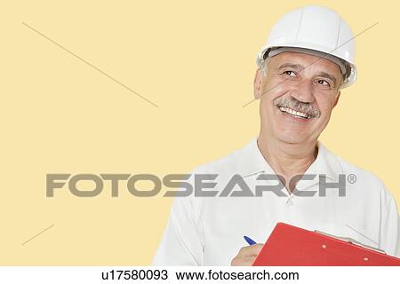 Stock Image - Senior constructor with clipboard looking away over yellow background. Fotosearch