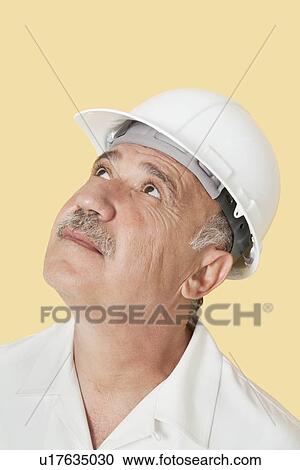 Stock Image - Senior man with hardhat looking up over yellow background. Fotosearch