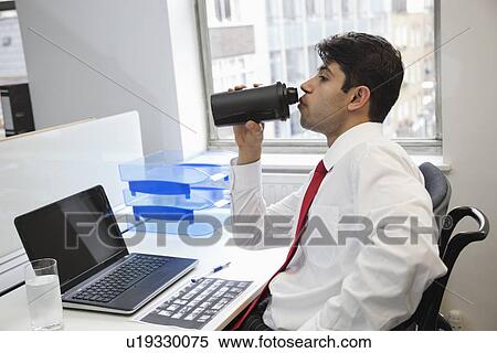 Stock Photography - Side view of an Indian businessman drinking water at office desk. Fotosearch