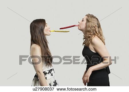 Stock Photo - Two young women blowing party puffers while looking at each other against gray background. Fotosearch