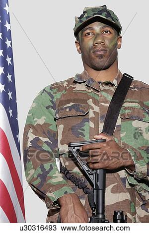 Stock Image - US Marine Corps soldier with M4 assault rifle standing by American flag over gray background. Fotosearch