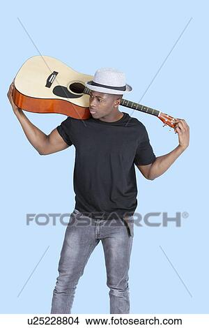 Picture - Young African American man carrying guitar over light blue background. Fotosearch