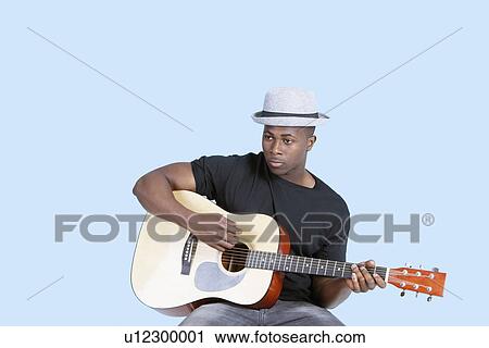 Stock Image - Young African American man playing guitar over light blue background. Fotosearch