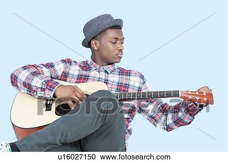 Stock Image - Young African American man tuning guitar over light blue background. Fotosearch