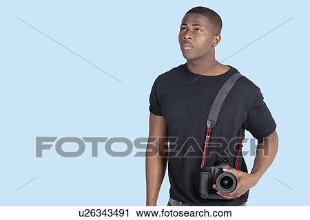 Stock Image - Young African American man with digital camera looking up over blue background. Fotosearch