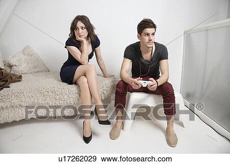 Young bored woman sitting on sofa with man playing video game View Large Photo Image Stock Photo - Young bored woman sitting on sofa with man playing video game. Fotosearch
